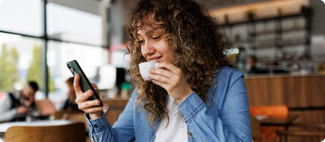 A woman sits in a café, smiling as she looks at her smartphone while holding a small cup of coffee.