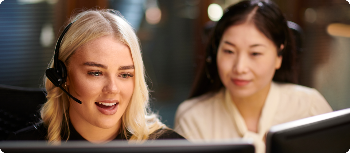 Two customer support representatives wearing headsets work at computer monitors in a modern office. One person in the foreground smiles while speaking, as a colleague looks on from behind.